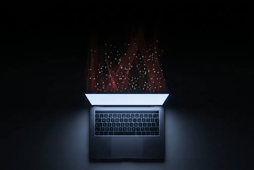 A laptop on a dark desk with red data threads rising from its glowing screen into darkness above